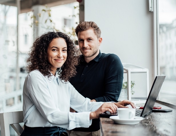 Ein Mann und eine Frau sitzen am Laptop und lächeln in die Kamera 