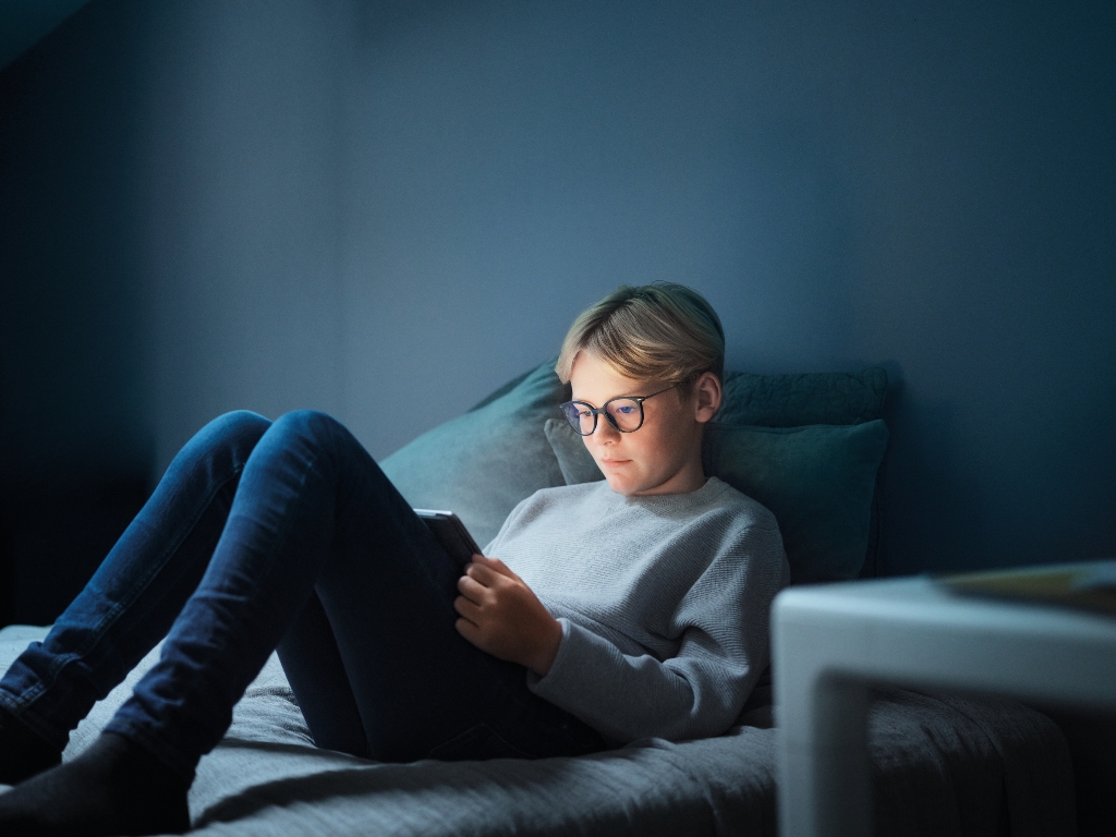 Junge mit Brille sitzt im Bett und schaut auf ein Tablet 